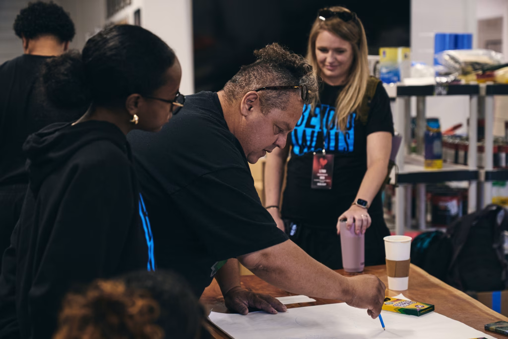 A group of people gather around a table as one person draws on paper with colored pencils. Two others watch closely, while another stands nearby smiling. Art supplies and drinks are visible on the table and shelves in the background.