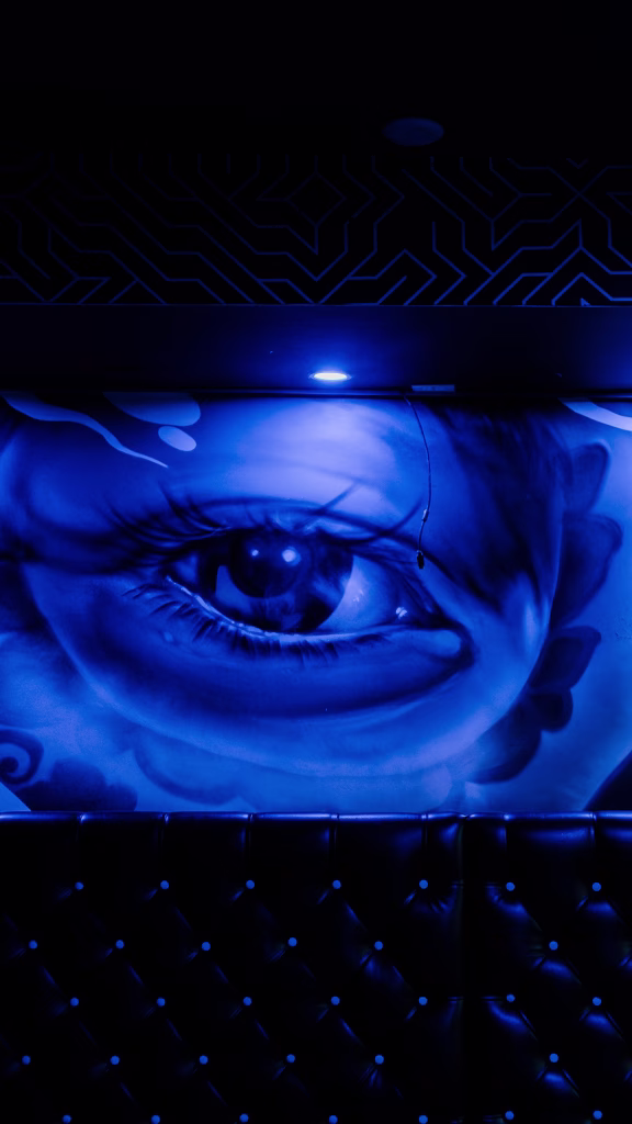 A large mural of a blue eye painted on a wall, illuminated by blue lighting. The lower part of the image shows a tufted black leather bench. The scene has a dramatic, moody atmosphere.