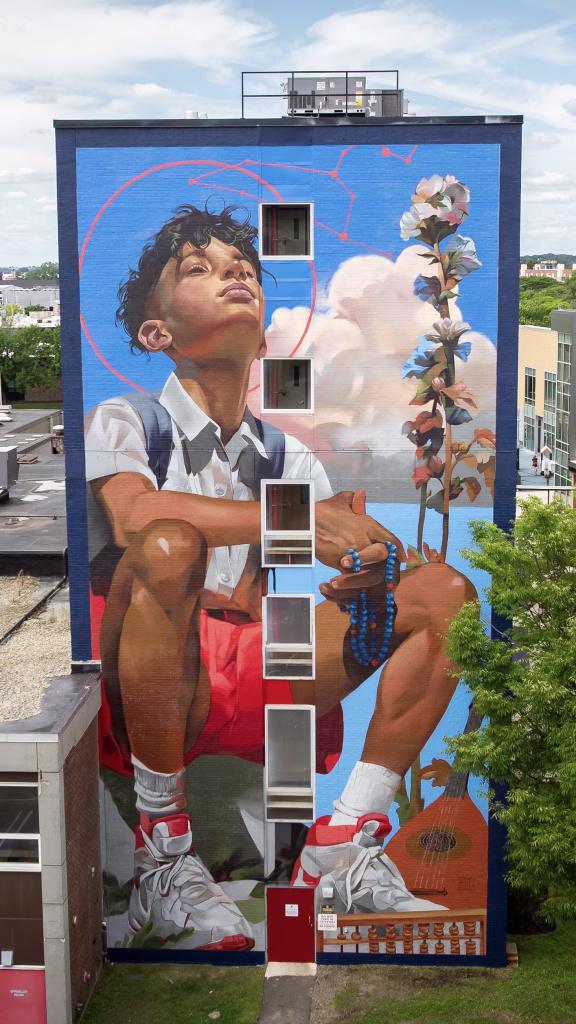 A mural on a building shows a boy in red shorts crouching, holding blue beads, with a flower and guitar beside him. He looks upward, eyes closed, against a vibrant blue sky and clouds.