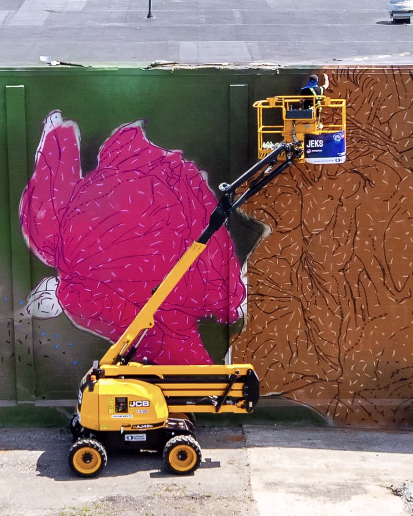 An artist uses a yellow cherry picker to paint a large mural of a pink flower with green stem and brown background on an outdoor wall.