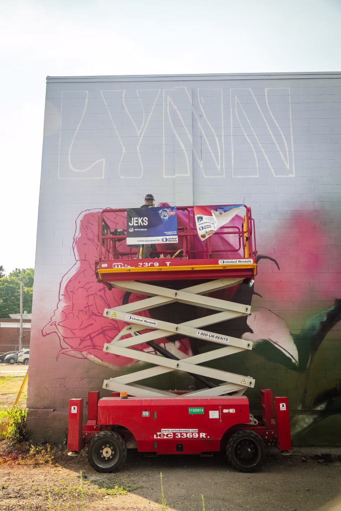 An artist on a red scissor lift paints a mural on a gray wall, outlining the word LYNN and a large floral design. Bright sunlight illuminates the scene.