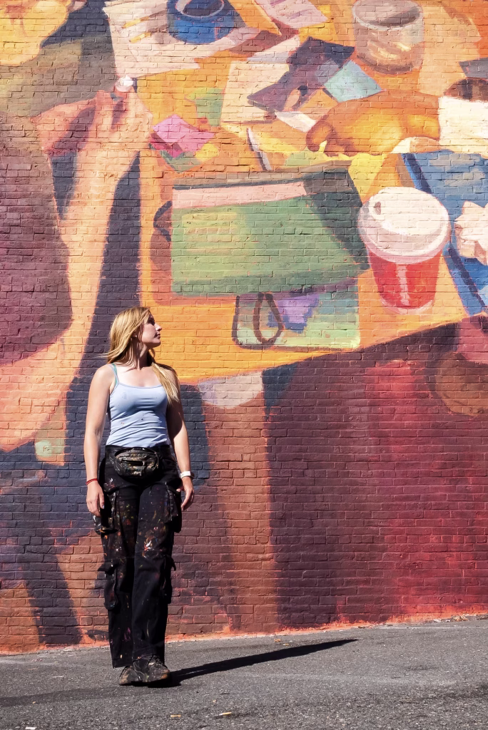 A woman in paint-splattered clothes stands in front of a colorful mural featuring a desk with scattered papers, notebooks, and cups, looking up at the artwork. Sunlight casts shadows on the wall.