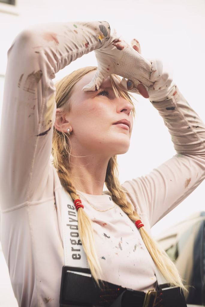 A woman with blonde braids, wearing a paint-splattered shirt and gloves, looks upward with one hand shading her eyes. She appears to be outdoors, possibly taking a break from painting.