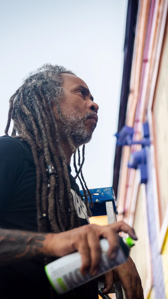 A person with long dreadlocks and a beard holds a spray paint can, standing near a mural in progress. The photo is taken from a low angle, with building details visible in the background.